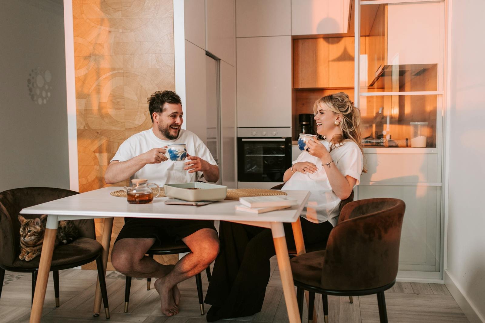 A happy pregnant couple sitting in a kitchen, enjoying tea and laughter together.